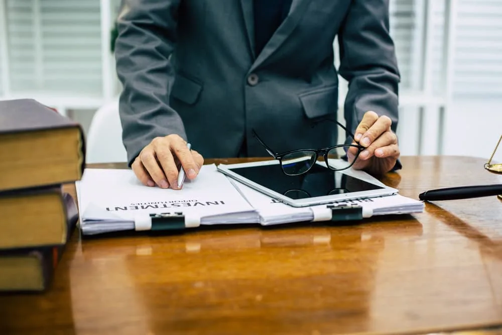 A person in a suit leans over a desk, holding a pair of glasses. There is a stack of books, a tablet, and folders labeled "Investment" on the desk.
