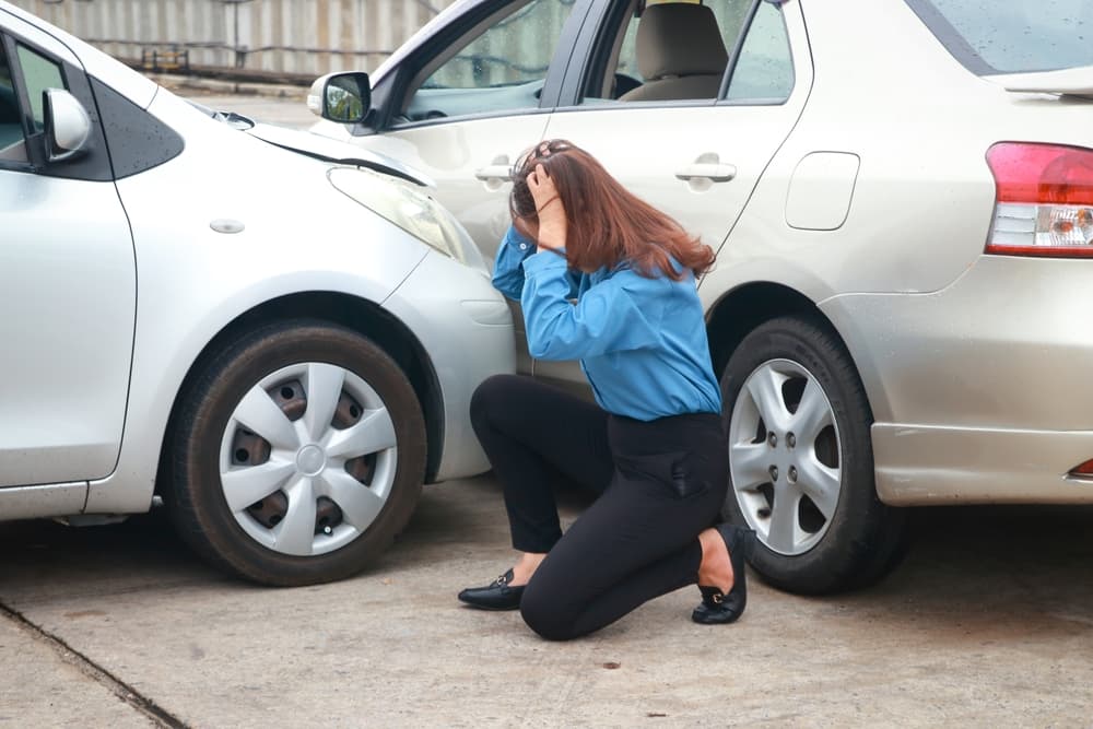 A stressed woman sits on the road, holding her head, gazing at the damaged cars after a collision.