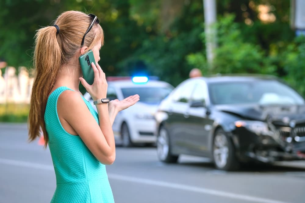 A stressed woman driver stands on the street side, talking on her mobile phone as she calls the police after a car accident.