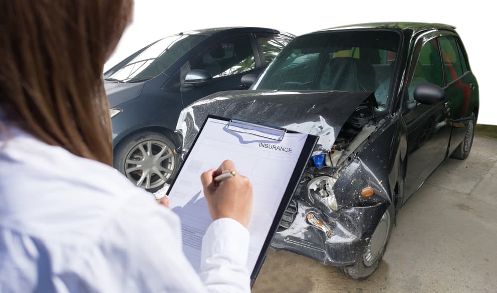 An insurance agent examines a damaged car while a customer signs the report claim form, part of the process after a traffic accident.