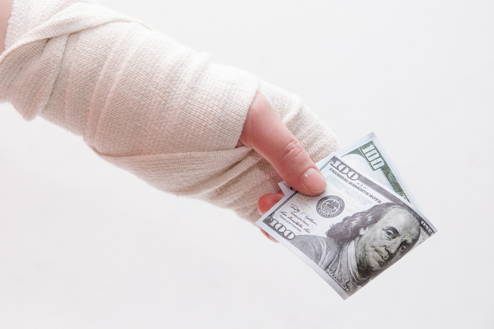 A female hand, wrapped in a bandage, holds a 100-dollar bill against a white background.