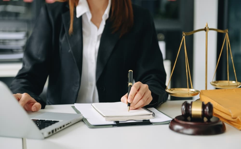  An woman lawyer works alongside a judge in a courtroom. The scene features a gavel, with the lawyer using a tablet, laptop, and digital tablet computer on a white surface.