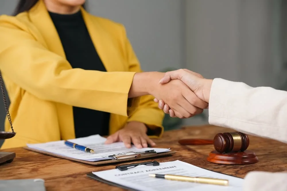 Two female lawyers shake hands after finalizing a contract agreement in an office, with legal documents and a gavel visible on the table.