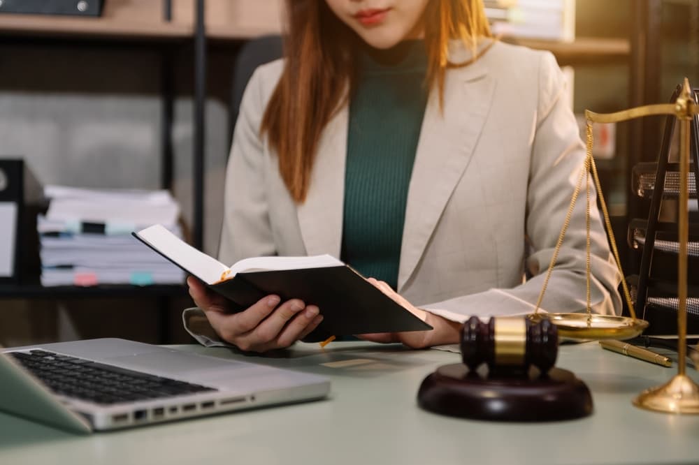 A woman lawyer reads a legal book with a gavel resting on the table in her office, symbolizing justice and law. 