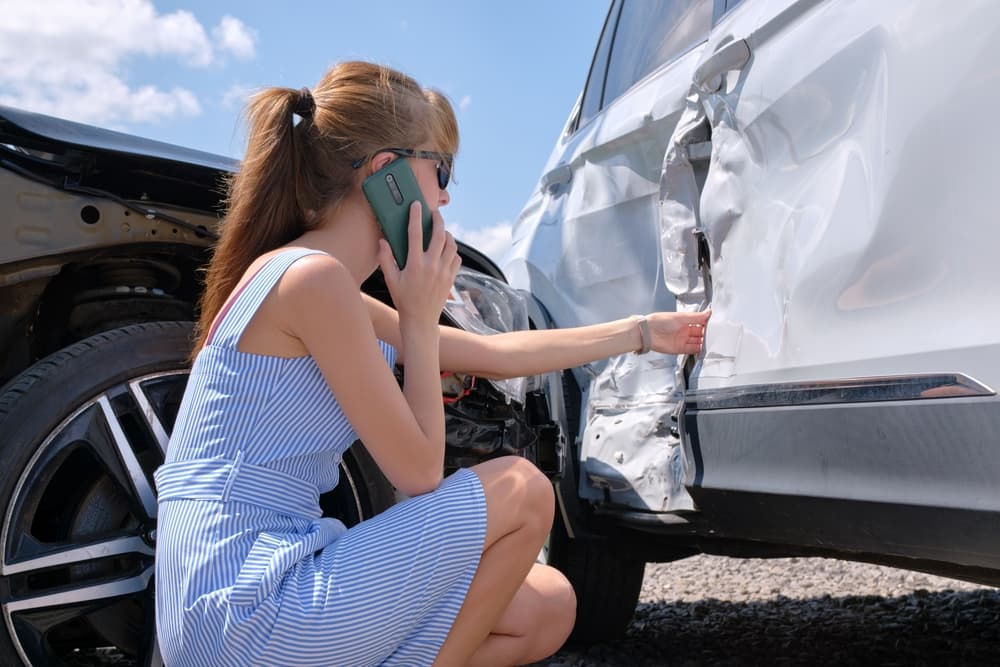 A stressed driver stands on the roadside near her smashed vehicle, talking on her cellphone as she calls for emergency service help after a car accident.