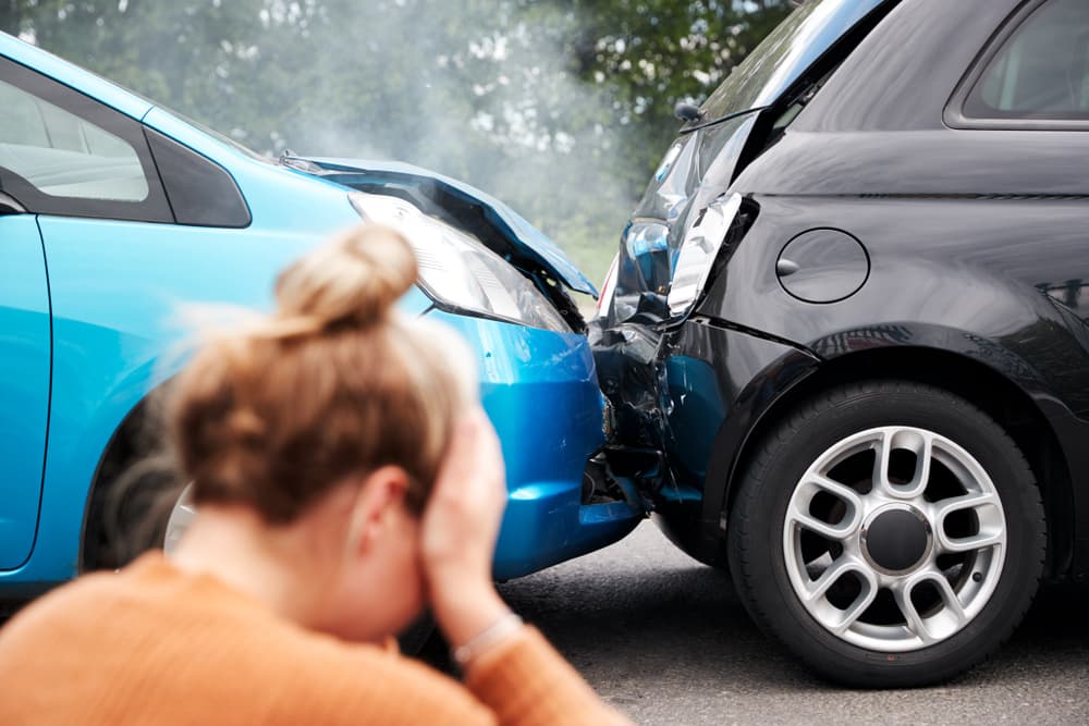 Female motorist sitting on the ground with her head in her hands, distressed, next to vehicles involved in a car accident.