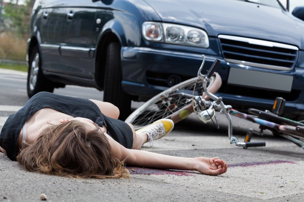 Unconscious female cyclist lying on the street after a road accident, with emergency responders and traffic in the background.