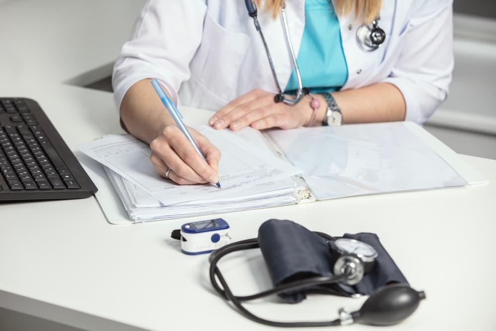 Close-up of a general practitioner in a white coat filling out paper medical records, writing notes, preparing documents, reports, and prescriptions at their workplace with a laptop in the background.