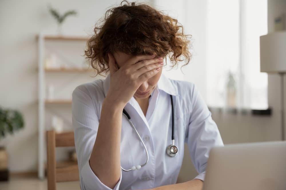 Exhausted and depressed young female doctor experiencing mental burnout at work, sitting alone at a hospital desk, feeling stressed and frustrated about medical failure and healthcare negligence, with her hand on her forehead in distress.