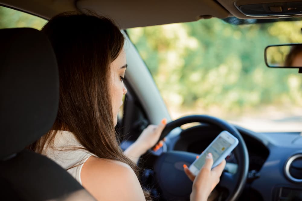 Young female student texting and writing messages while driving a car, not paying attention to the road, illustrating dangerous driving and social media addiction.