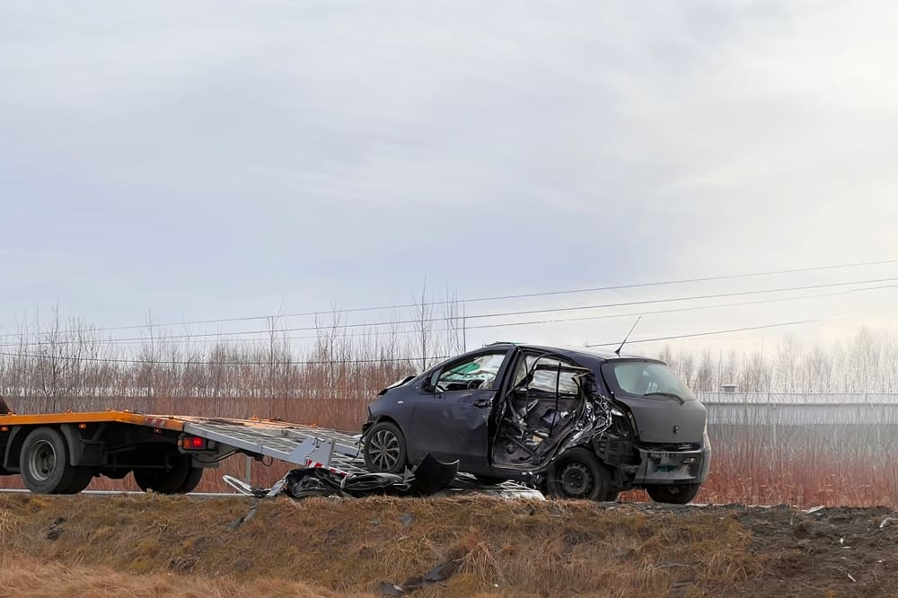 T-bone road accident with a modern family car severely damaged on its side. A tow truck transports the broken car on the highway, marking the accident site.