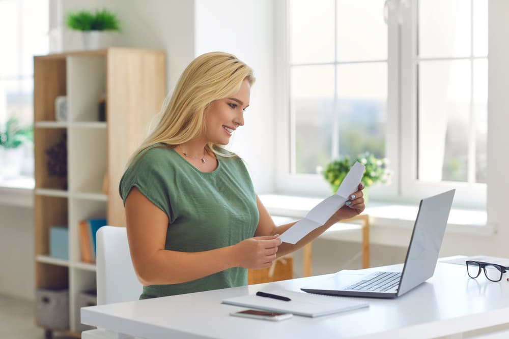 Happy young woman sitting at a desk at home, reading a document about insurance approval or receiving a money refund.