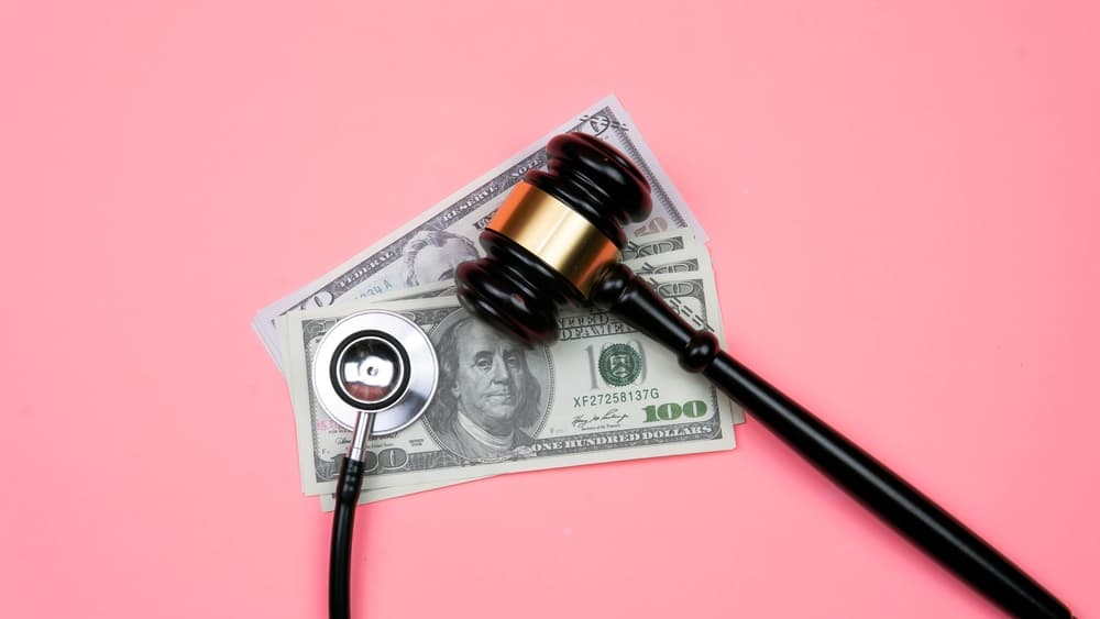 Wooden gavel, a stethoscope, and U.S. dollar bills on a pink background.