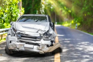 A silver car with severe front-end damage is parked on the side of a road bordered by trees on a sunny day.