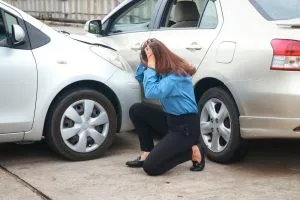 A woman kneels on the ground holding her head in distress next to two cars involved in a minor collision, with visible damage to both vehicles.