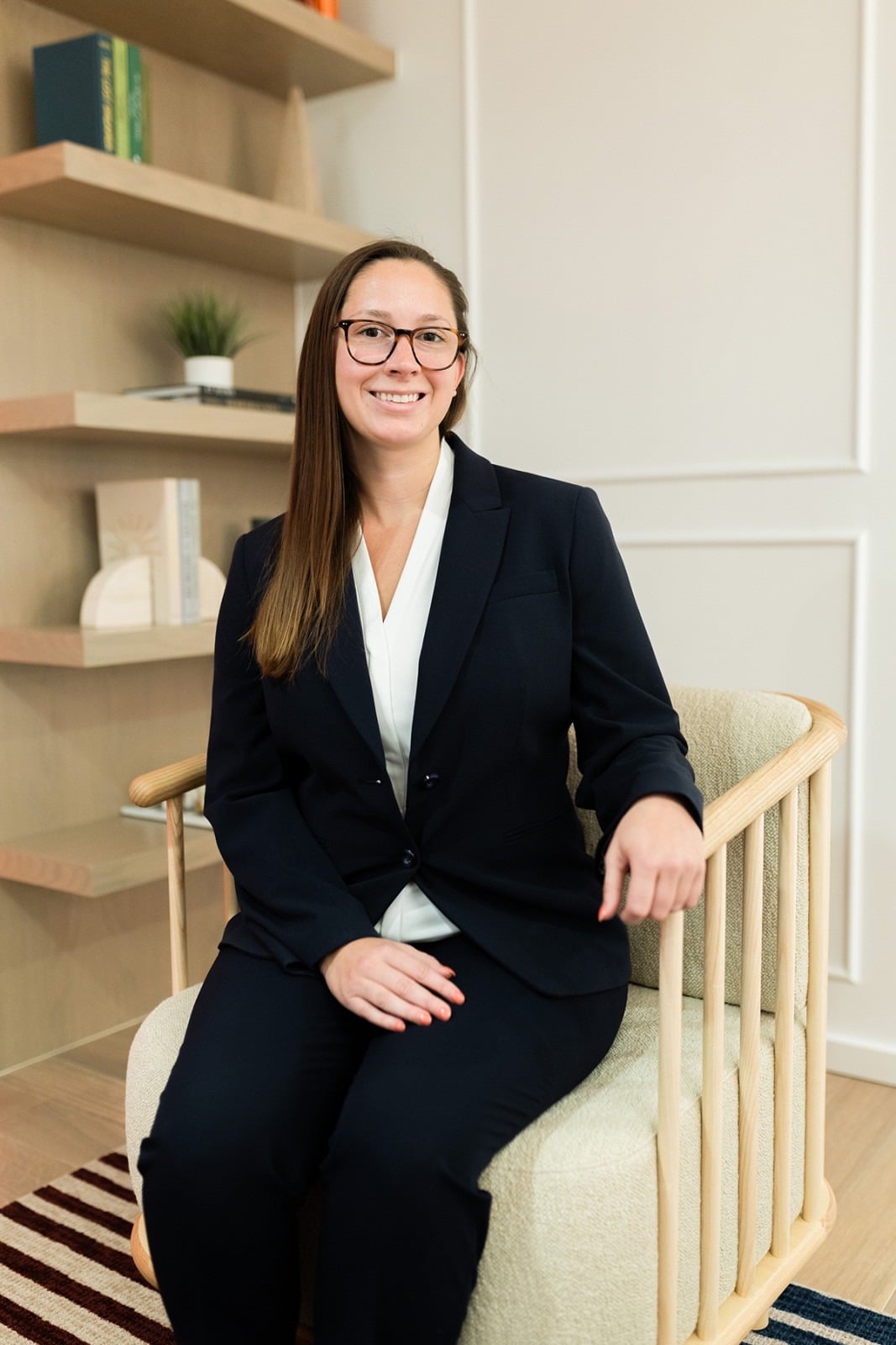 A woman in a dark suit and glasses sits on a chair in a modern office space with bookshelves in the background.
