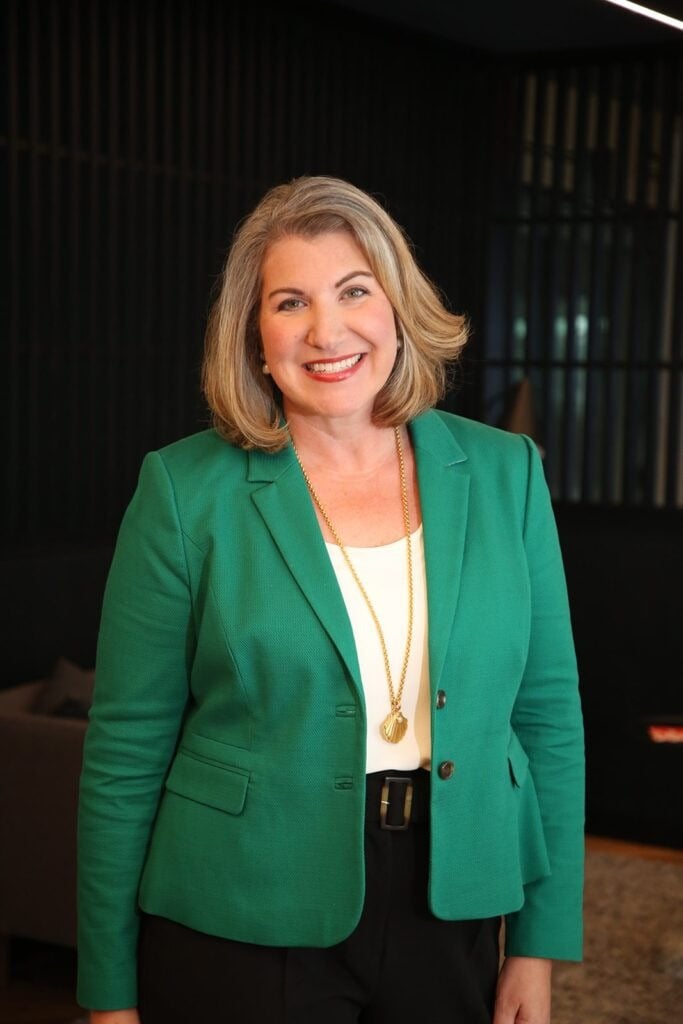 Woman with shoulder-length blonde hair smiles and stands indoors, wearing a green blazer, white top, black pants, and a long gold necklace.