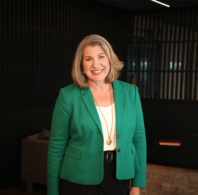 A woman with shoulder-length gray hair, wearing a green blazer and white top, stands indoors and smiles at the camera.