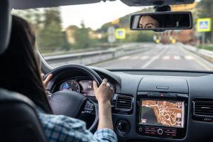 Person driving a car, hands on the steering wheel, with a navigation map displayed on the dashboard screen and road signs visible through the windshield.
