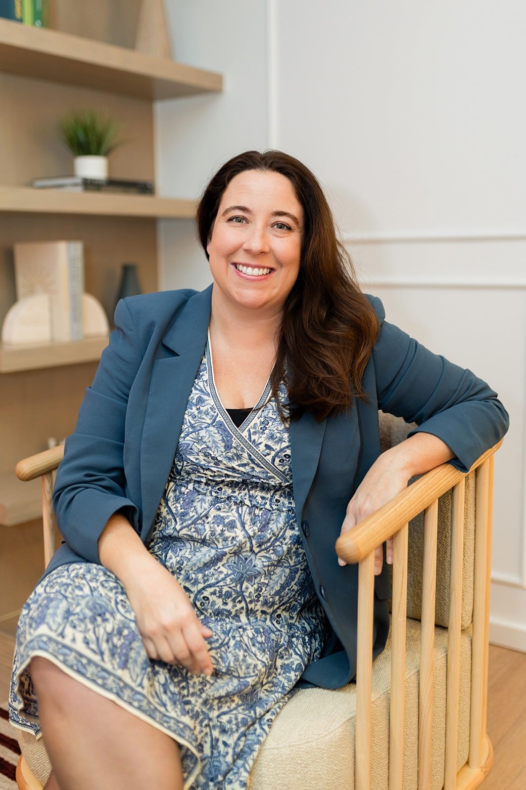 A woman with long brown hair, wearing a blue patterned dress and blue blazer, sits and smiles in a wooden chair in a modern, well-lit room with shelves behind her.