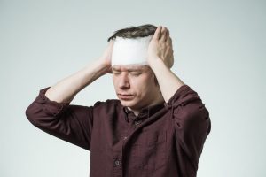 A man with a bandaged forehead holds his head with both hands and closes his eyes, appearing to be in pain or discomfort.
