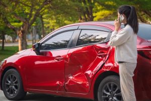 A woman stands by a red car with a large dent in the driver's side door, talking on her phone, in an outdoor setting with trees in the background.