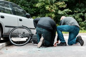 Two people kneeling beside a fallen bicycle next to a parked car on a street, with broken glass scattered on the ground.