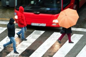Three people cross a wet street at a crosswalk; one holds an orange umbrella, two wear hooded jackets. A red bus is visible in the background.