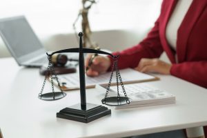 A balance scale is on a desk with legal documents, a gavel, and a person in a red blazer working in the background.