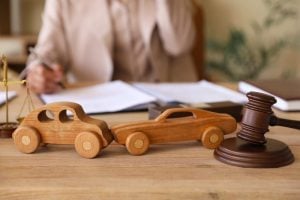 Two wooden toy cars are positioned as if in a collision on a desk, with a judge's gavel and legal documents in the background.