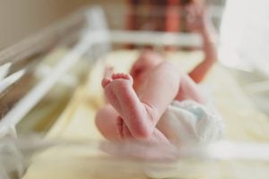 A newborn baby lies on its back in a hospital bassinet, wearing a diaper, with feet in focus and legs slightly raised.