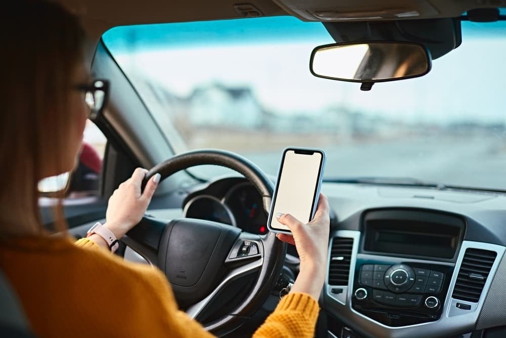 A person in a yellow sweater sits in a car, holding the steering wheel with one hand and using a smartphone with the other. The phone screen is blank.