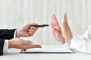 One person offers a pen and paper for signing while another person refuses with both hands raised in a stop gesture.