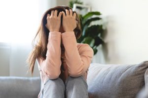 A person sits on a couch with their knees up, covering their face with both hands, appearing distressed. Indoor setting with a plant in the background.