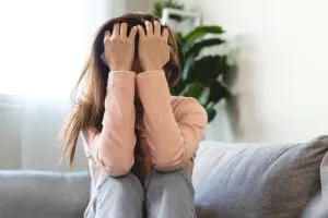 A person sits on a couch with their knees up, covering their face with both hands, appearing distressed. Indoor setting with a plant in the background.