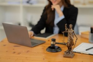 A woman in business attire works on a laptop at a desk with a gavel and a Lady Justice statue in the foreground.