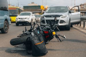 A motorcycle lies on its side next to a backpack on a road, with a car door open nearby and traffic stopped in the background.