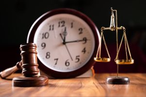 A wooden gavel, a brass balance scale, and a clock are placed on a wooden surface, symbolizing justice and the passage of time.