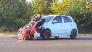 A red motorcycle is crashed into the front of a white car on a road, with trees and greenery in the background.