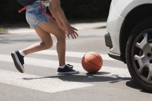 A person reaching for a basketball on a crosswalk near a white car.