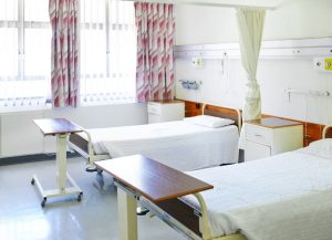 A hospital room with two empty beds, bedside tables, medical equipment on the wall, and sunlight coming through curtained windows.