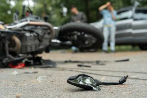 A damaged motorcycle and broken glass lie on the road after a crash, with two people standing near a car in the background.
