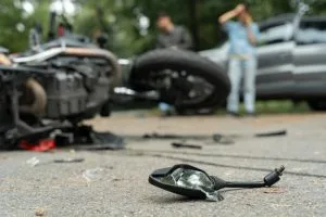 A damaged motorcycle and broken glass lie on the road after a crash, with two people standing near a car in the background.