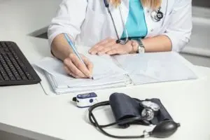A healthcare professional in a white coat writes on a clipboard at a desk with a keyboard, blood pressure monitor, and pulse oximeter.