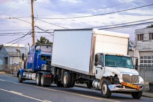 A damaged white box truck is being towed by a blue semi-truck on a city street, with industrial buildings visible in the background.