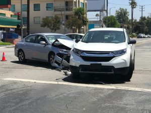 Two cars, a silver sedan and a white SUV, are involved in a front-end collision at an intersection on a sunny day.
