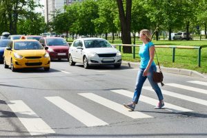 A woman in blue jeans and a turquoise shirt crosses a city street at a crosswalk, with several cars stopped in the background.