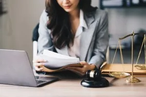 A woman in a suit sits at a desk with a laptop, reviewing documents; a gavel and scales of justice are also on the desk.