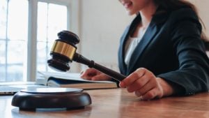 A person in a suit holds a judge’s gavel at a desk with an open book and a laptop in front of them.