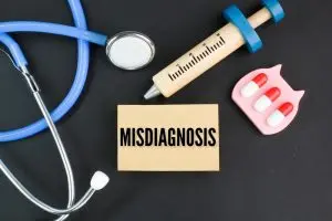 A stethoscope, syringe, and three pills surround a card labeled "MISDIAGNOSIS" on a black background.